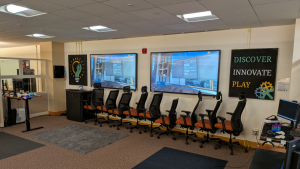 A room with rugs spread out and a row of chairs alongside two monitors displaying a VR experience.