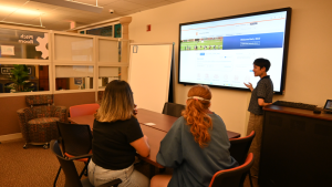 A student presenting to two other students in the pitch room, which includes eight rolling chairs, two six foot narrow tables joined together, and an instructor station connected to a monitor on a wall.