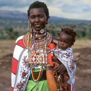 A Maasai woman wearing multi-colored, beaded jewelry holds a child on her hip.