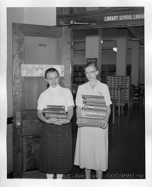 Two library students stand just outside of the Library School Library, holding stacks of books.