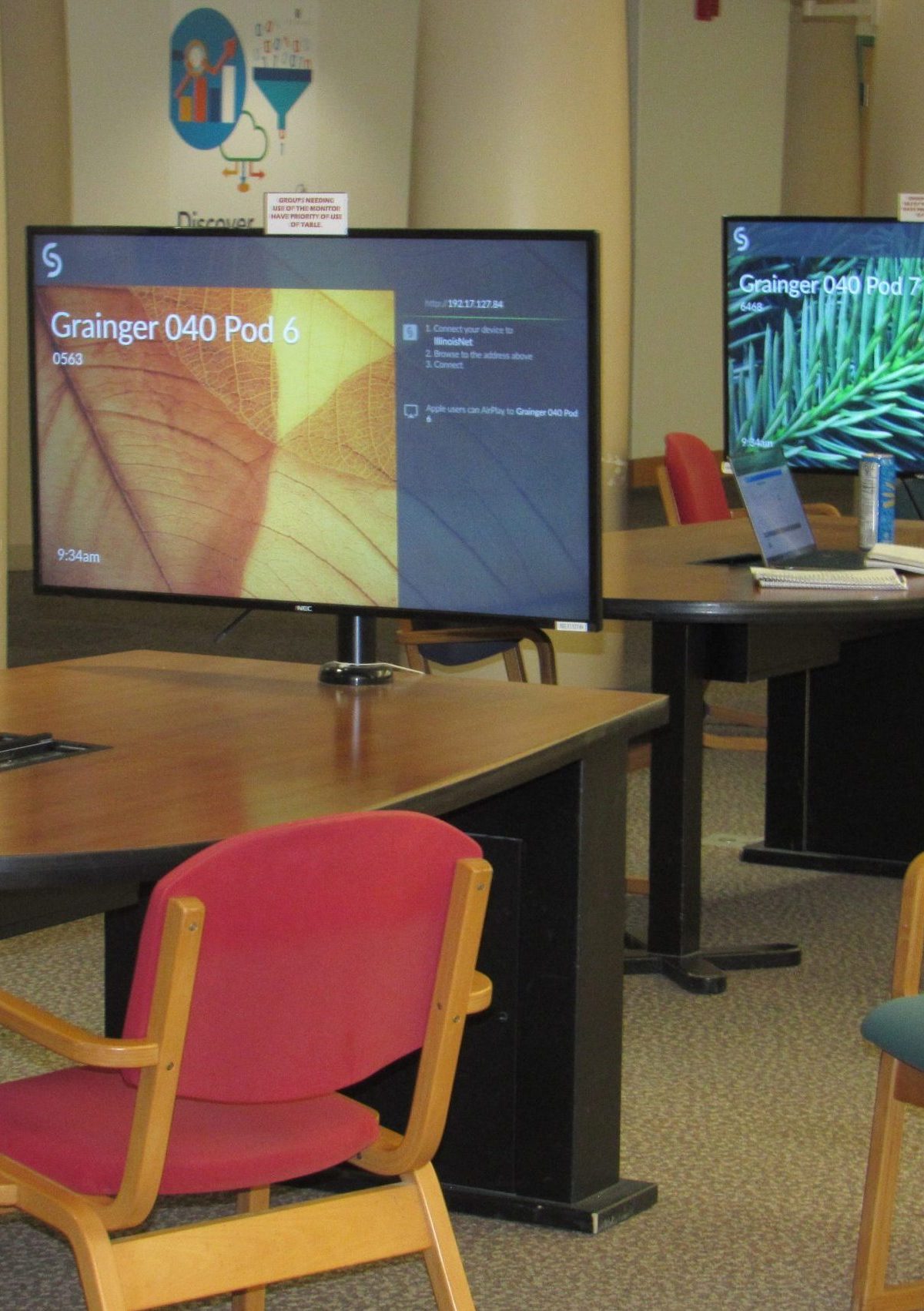 Basement Study area in Grainger engineering library with round wooden tables and chairs in red and blue. Each table has a large monitor displaying 'Grainger 040 Pod 6' and 'Grainger 040 Pod 7' with images of leaves on the screens. Some tables have laptops, notebooks, and drinks. The room has carpeted floors