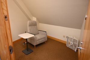 A room with an armchair with a small table next to it. There is a box fan in the corner.