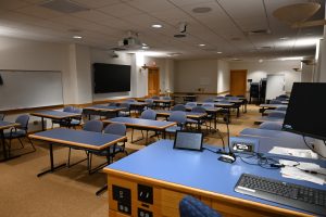 A large room with multiple tables and chairs. A desk with a computer is at the front. There are television screens around the room.