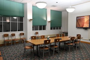 A room with a long table with chairs surrounding it. There is a television screen on the wall and three large windows.