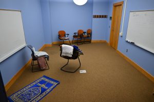 A room with multiple chairs. There is a whiteboard on one wall. There is a prayer mat and other religious objects throughout the room.