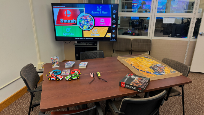 A gaming set-up featuring a monitor, multiple chairs, and two joined tables. The tables have a puzzle, board game, and an assortment of Nintendo Switch games. The monitor is displaying the Nintendo Switch game Smash Bros.