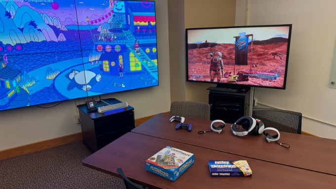 A gaming set-up featuring one large monitor displaying a lofi Pokemon graphic while another smaller monitor shows gameplay from the PS5 game No Man's Sky. Multiple chairs and two joined tables are in the room displaying PlayStation 5 controllers, PSVR2 headset, and board games.