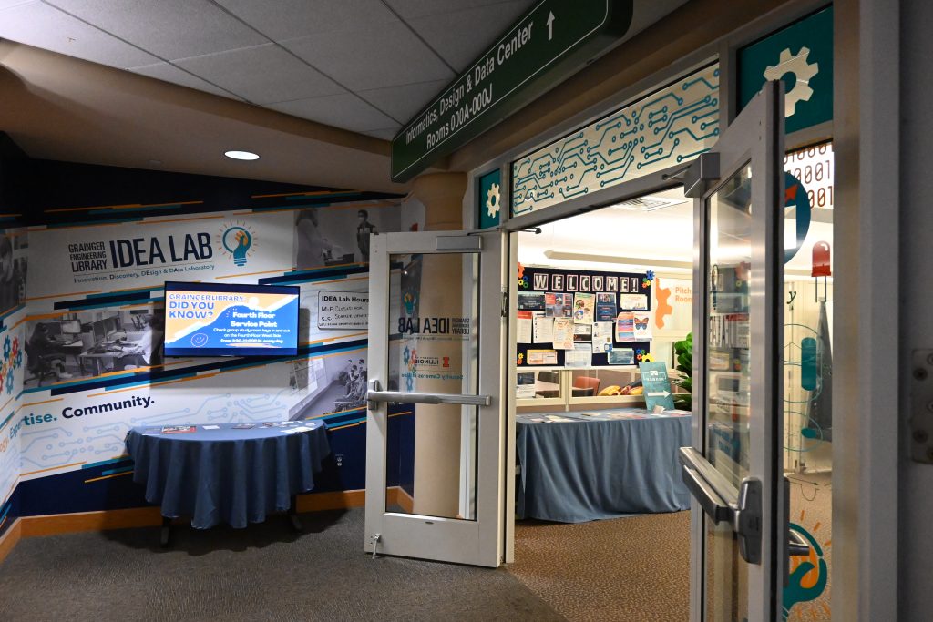 Entryway of the IDEA Lab featuring a monitor above a side table, the double doors of the lab entrance, and a welcome sign. Cubicles and a bulletin board are further in the lab's space.