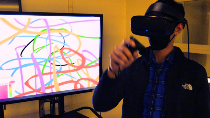 A person wearing a VR headset and using VR controllers to virtually paint with a monitor displaying paint strokes in a VR canvas