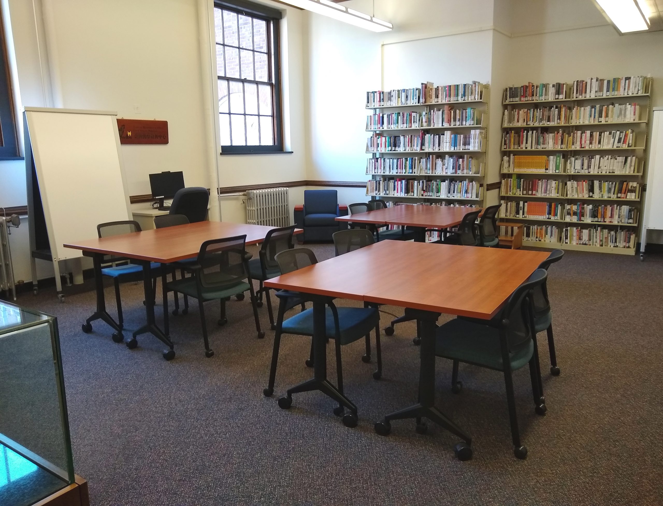 Three tables with chairs in a mid-sized, carpeted room with bookshelves and windows in the back.