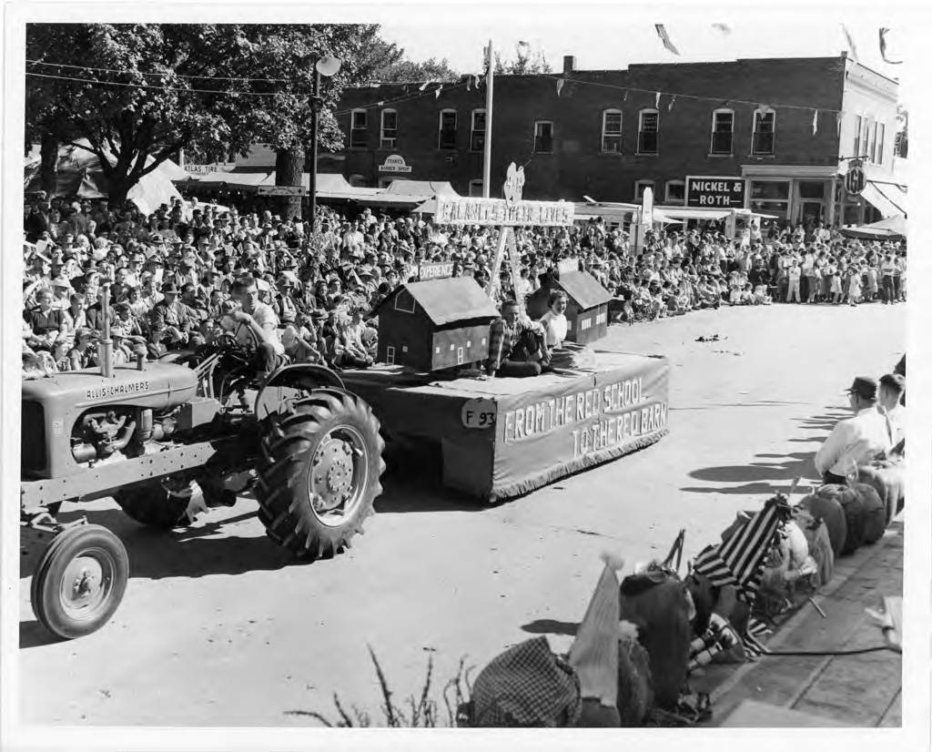 A tractor driven by a man in glasses is pulling a parade float with two people sitting on it. On either side of the people is a small house. On the front of the float is says "From the red school to the red barn." There are hundreds of people lining the streets of the parade.