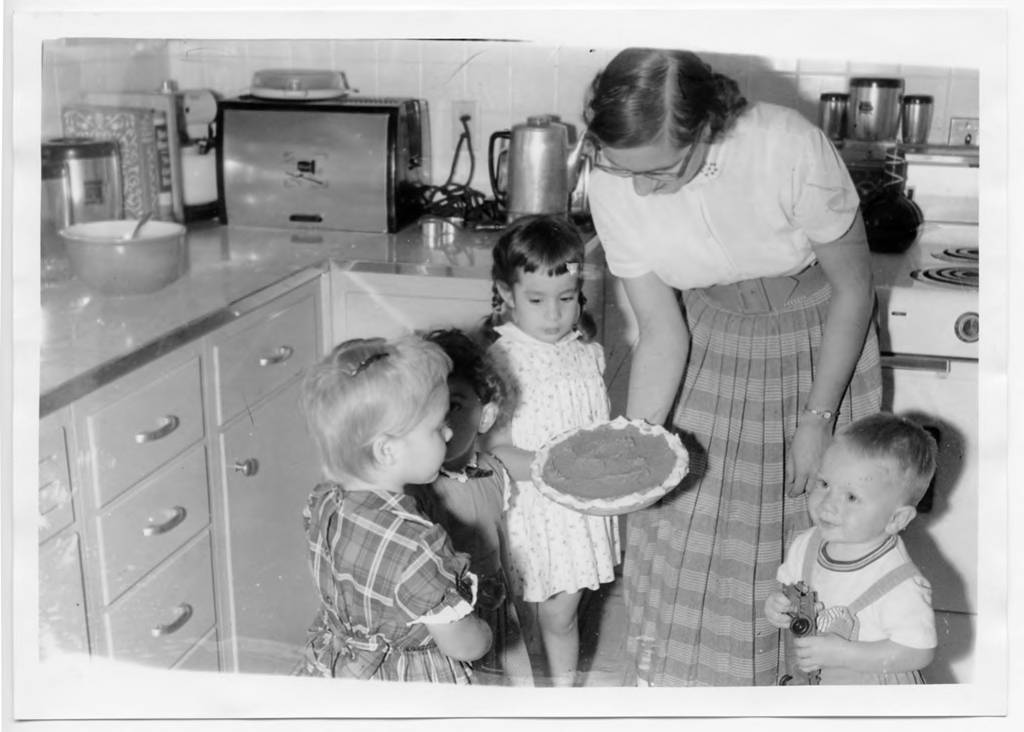 A woman and four children look at a freshly baked pie. They are standing in a kitchen.