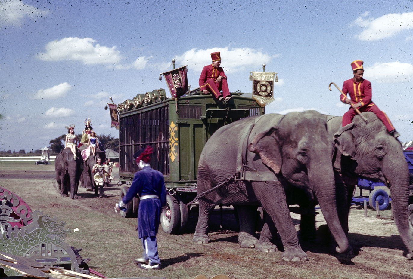 Color image of two elephants pulling green circus cage wagon. Men in red outfits sit atop one elephant and the cage wagon.