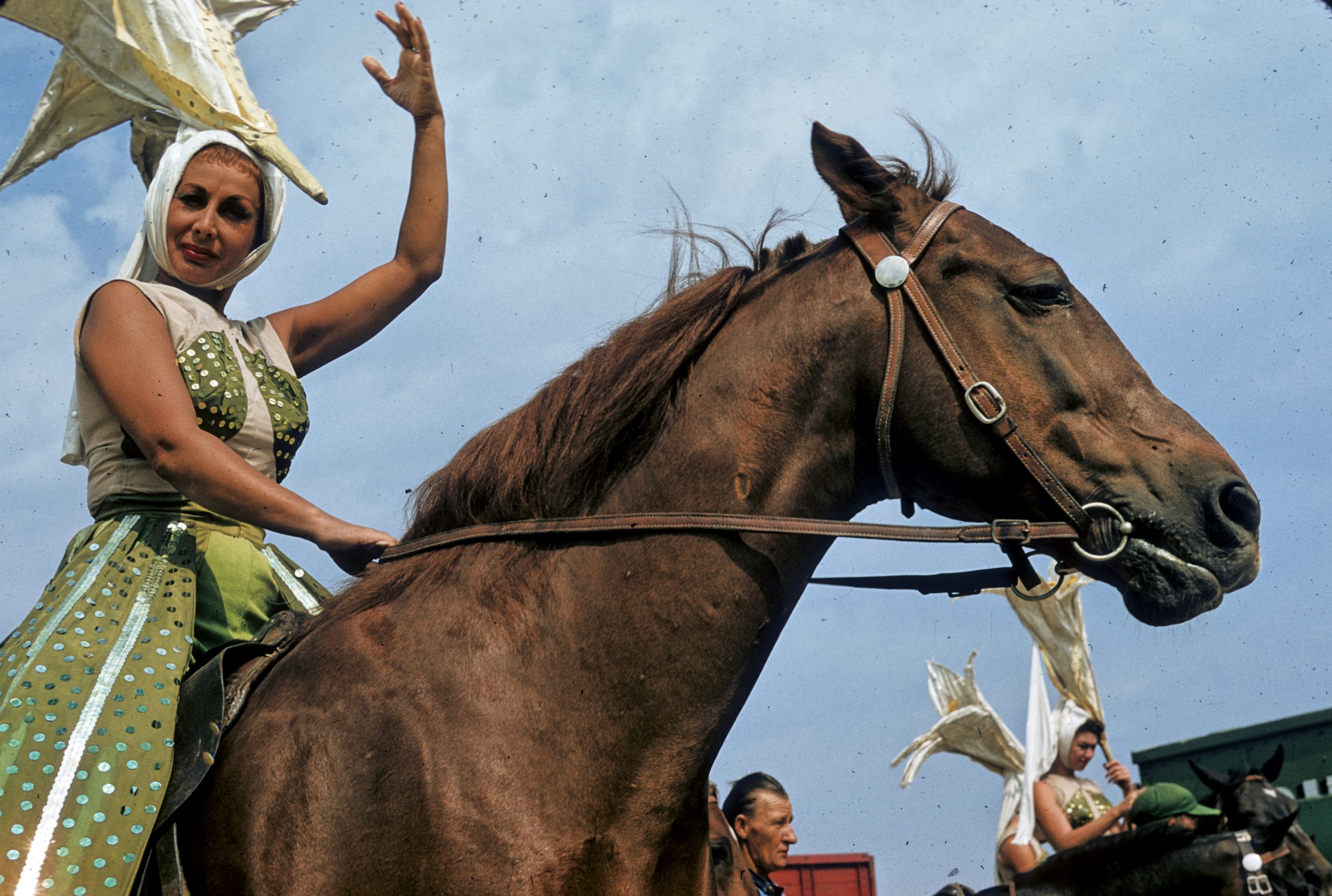 Image of a woman wearing a large star headdress and green and white outfit, extending her arm and sitting on top of a horse.