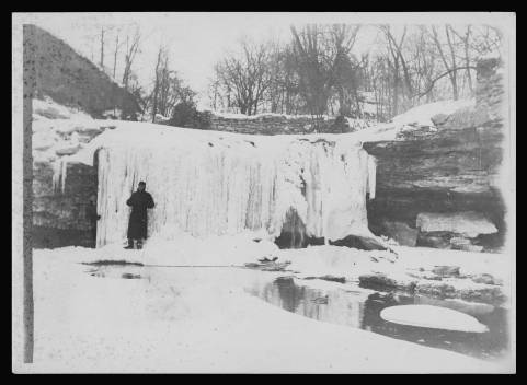 Black and white photo of a person standing in front of a frozen waterfall