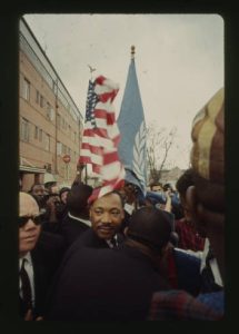 Martin Luther King, Jr. is in a crowd, an American flag flies above him