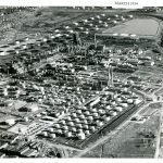 Black and white aerial photograph of refinery tanks and stills along railroad track, with some homes in background. Top right corner labeled "MARCH 1954"
