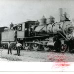 Two men pose in front of a locomotive connected to a coal car and box car