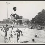 Black and white photo of people at a pool. A lifeguard sits on a platform in the foreground.