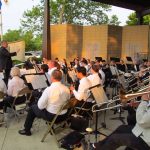 An orchestral band including brass instruments, woodwinds, and percussion, performs outside in a band shell