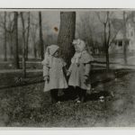 Black and white photo of Marcelline and Ernest Hemingway as very young children dressed in winter attire and holding hands in front of a large tree.