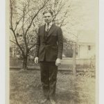 Black and white photograph of Ernest Hemingway as a teenager dressed in a three-piece suit and standing in front of a small tree and fence.