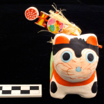 One piece molded and painted cat figure. Painted red, white and green. Has wooden toy and silk rattle on back