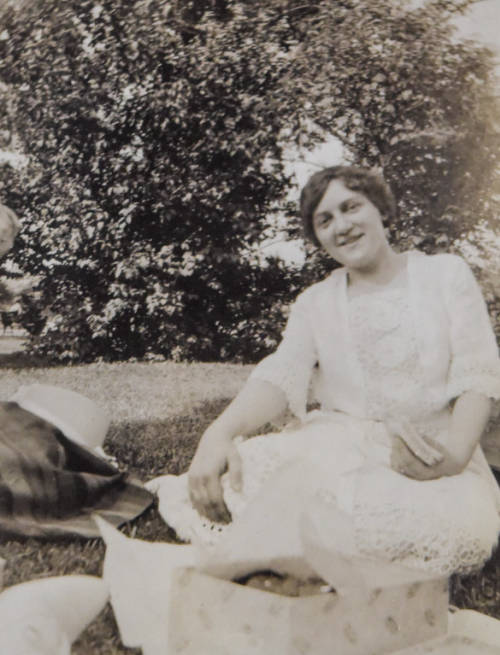 Woman holding a sandwich while sitting on the ground. Food and a picnic blanket are around her.