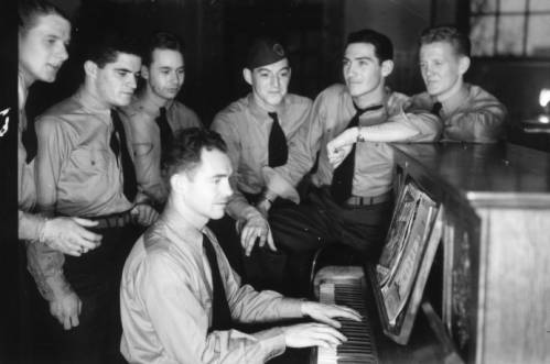 A group of naval flight school cadets gather around an upright piano as one cadet plays. 
