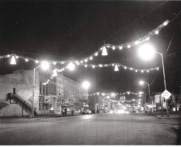 black and white photo of Christmas lights and wreaths hung on wires crossing over a history downtown street