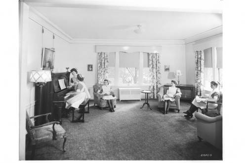 A group of nurses on a break sit on chairs in an open room while another nurse plays a piano