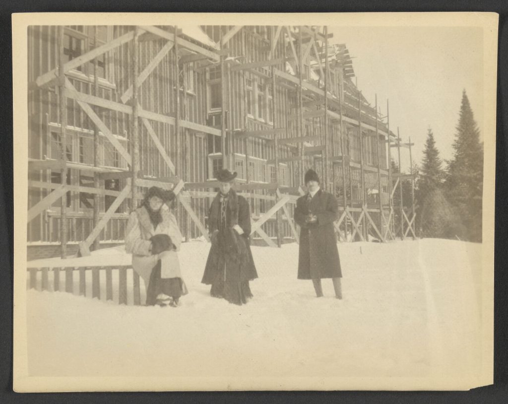 black and white photograph of three people standing in front of a house construction frame on a snowy day.
