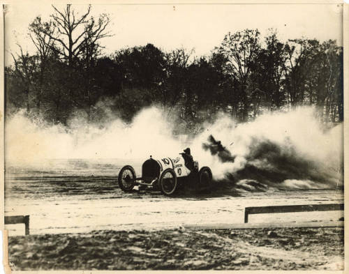 sepia-toned photo of a white racing car from the early twentieth century in a large dust cloud on a dirt race track.
