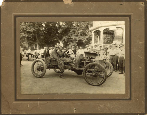 sepia-tone photo of two men in coveralls and dustcaps sitting in an open racing car from the early twentieth century.