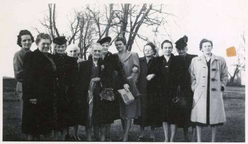 black and white photo of a group of women, mostly elderly, wearing coats and standing outside in front of bare trees.