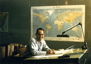 James A. Bier in November 1957, sitting at a drafting table with a map of the world posted on the wall behind him.