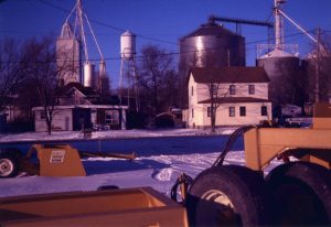 Gridley, IL, in winter showing farm equipment in the foreground, assorted single-family homes in the mid-ground, and multiple grain elevators and elevated water tower in the background.