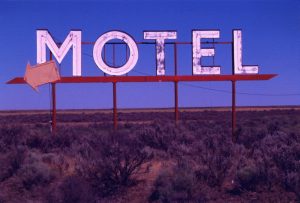 Large roadside Motel sign. Letters are white with neon tubing for nighttime illumination and are mounted on red frame elevating them above arid landscape of bunchgrass and sagebrush. Sign includes red arrow, also with neon tubing, pointing toward the left.