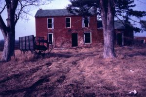 Abandoned brick farmhouse near Henning, IL, with pair of trees, one appearing dead, in front. House windows and doors are missing; unidentified farm equipment is underneath dead tree to the left. Grass is brown and not cut; earth is rutted by vehicles.