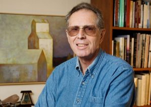 Professor Emeritus John A. Jakle in his study wearing a blue denim shirt and posed in front of an impressionistic painting of grain elevators and a bookshelf.