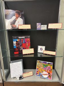 Exhibit case featuring vinyl records, cassette tapes, CDs, and printed music. The items included represent popular hits, new media formats (cassettes, then CDs), and musical compositions with accompanying media. 