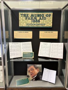 Vertical exhibit case with information about farms in crisis, the first Farm Aid concert in 1985, scores open to the music for songs by John Mellencamp and others, an album by Johnny Cash, and a book open to the lyrics of Folsom Prison Blues. 