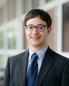 An image of a young white male in a business suit and glasses, smiling at the camera.