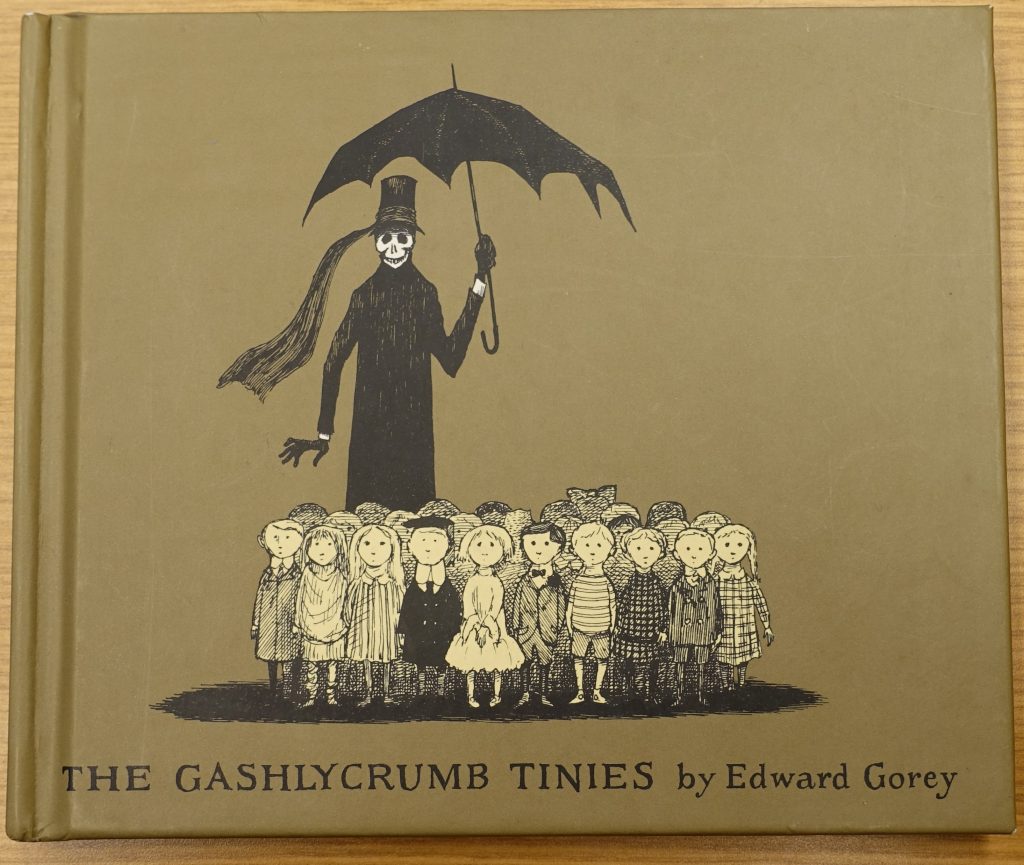 The front cover of the book "The Gashlycrumb Tinies," which shows a tall figure in black with a skull for a face holding an umbrella over a group of children.