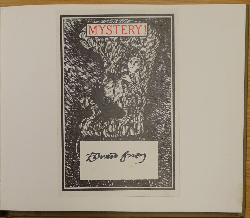 A bookplate signed by Edward Gorey. It shows a man sitting in a high-backed armchair reading a book with a black cat at his feet. It reads "Mystery!" in red at the top.