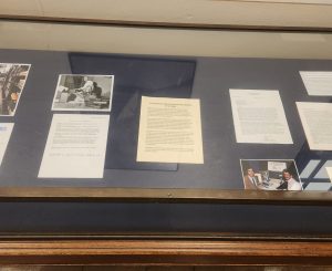 Shows documents and photos resting on a blue felt backing in a glass, metal, and wood exhibit case.