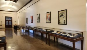 Angled photo showing large, high-ceiling room with five framed posters and five glass, wood, and metal exhibit cases.